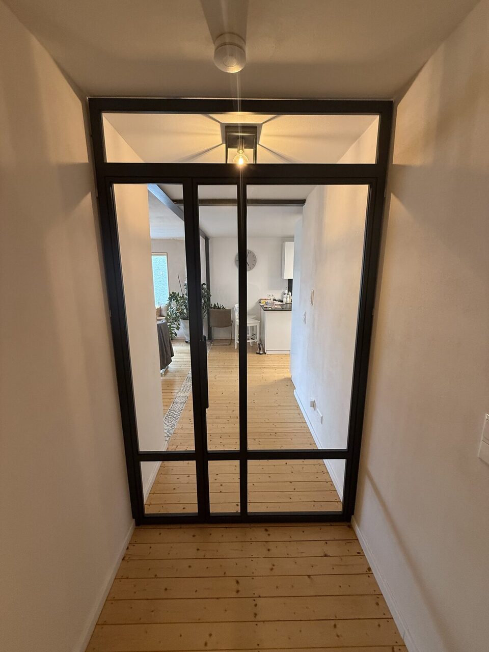 Korridorperspektive durch eine Stahl-Glas-Lofttür in ein helles Wohnzimmer