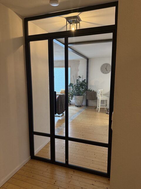 Stahl-Glas-Lofttür frontal vor einem hellen Wohnzimmer mit Fensterfront
