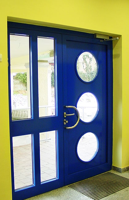 Blaue Metalleingangstür mit runden Glasausschnitten vor gelber Wand